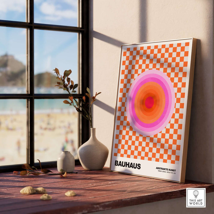 Bauhaus art print featuring pink circles on an orange checkerboard, displayed on a windowsill with a beach view.