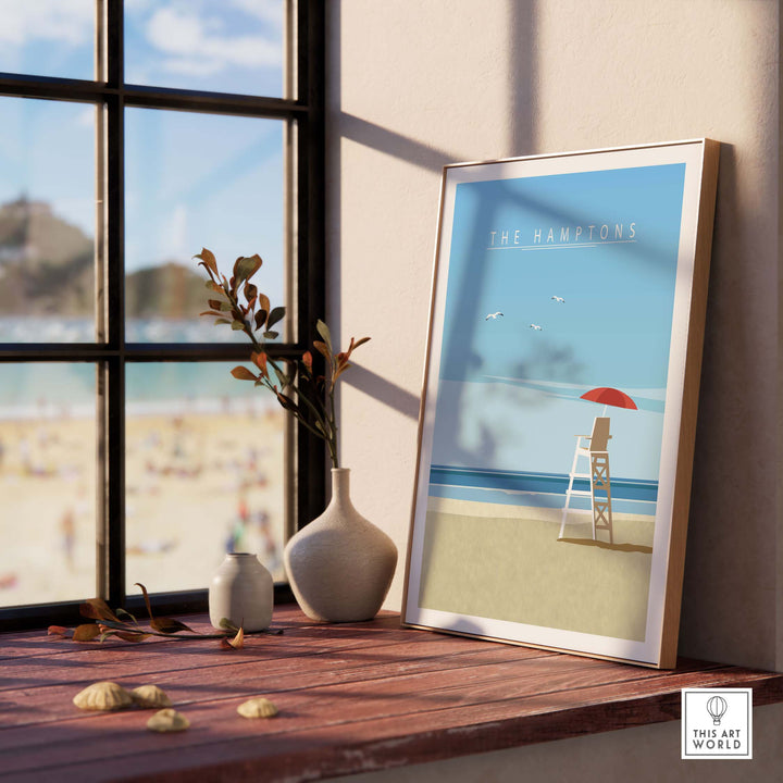 A framed travel poster print titled 'The Hamptons' displayed on a windowsill with beach view, capturing minimalist summer vibes.