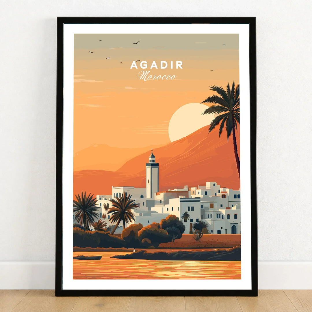 Agadir Travel Poster – Moroccan Mosque and Desert Sunset Art in a black wooden frame leaning against a white wall on a light wooden floor
