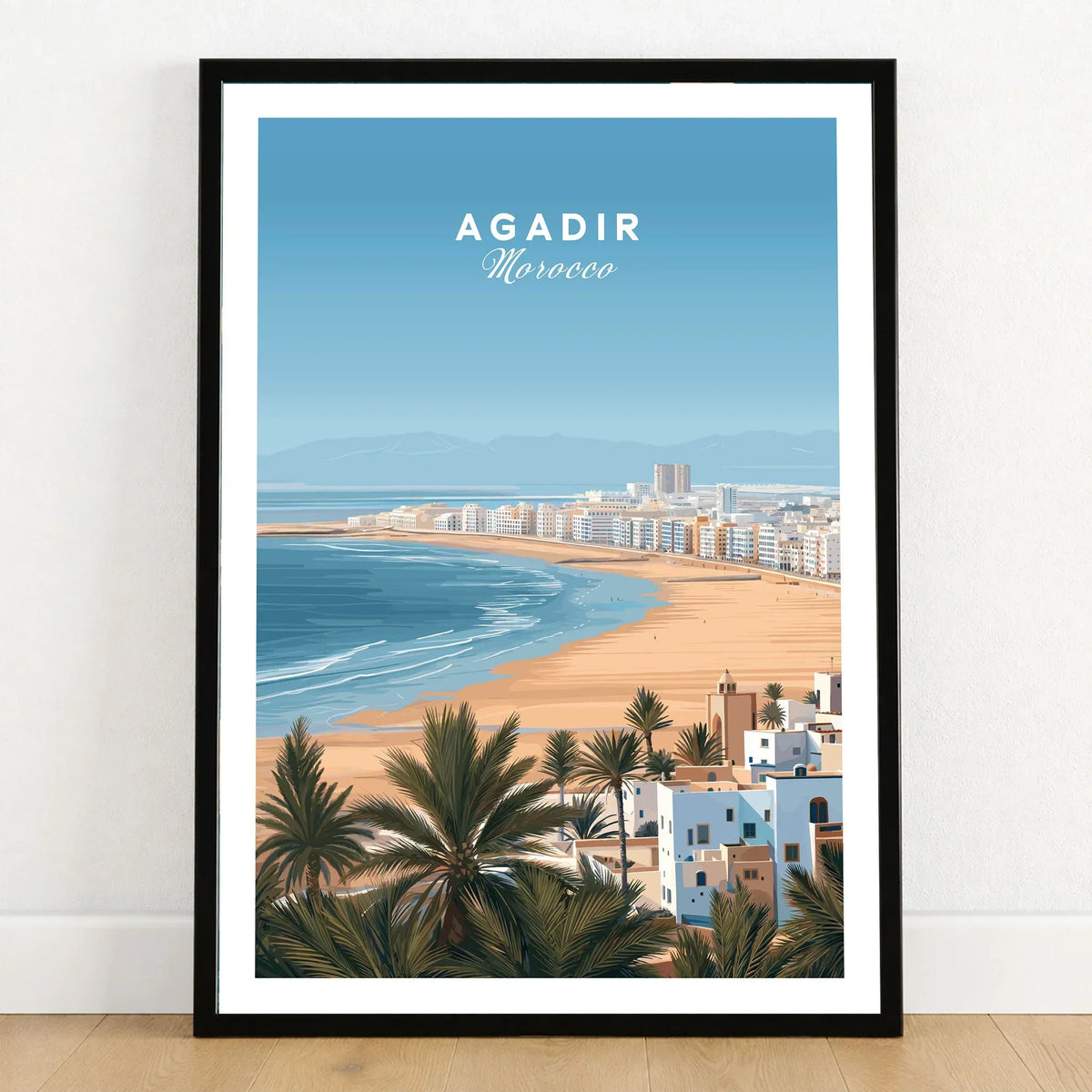 Agadir Morocco Poster – Travel Print in a black wooden frame leaning against a white wall on a light wooden floor