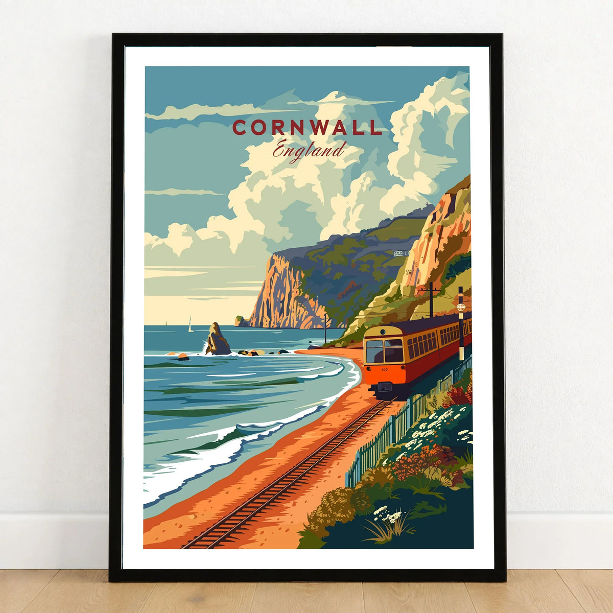 Cornwall Coast Travel Print in a black wooden frame leaning against a white wall on a light wooden floor