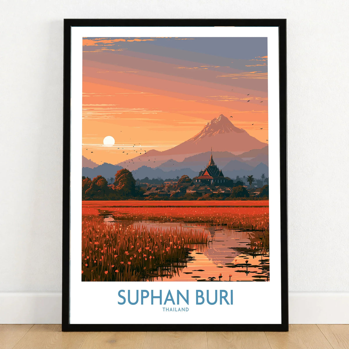 Suphan Buri Thailand Temple Sunset Wall Art — a lotus-covered lake and temple spire at sunset beneath a mountain peak, shown as art print in black frame on wooden floor