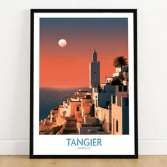 Tangier Morocco Coastal Sunset Wall Art — a whitewashed hillside town and minaret glowing above the sea at sunset, shown as art print in black frame on wooden floor