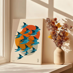 Modern canvas wall art with blue birds and orange sun, next to vase of dried flowers by sunny window