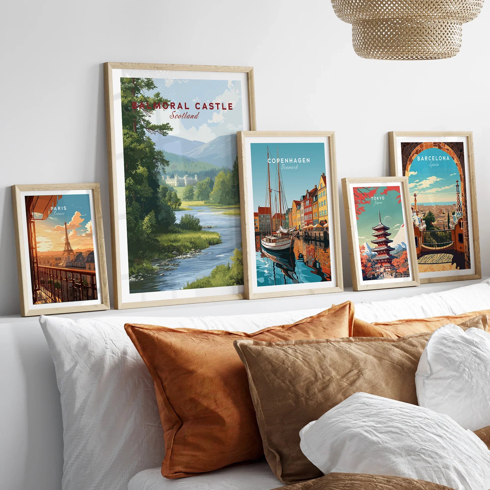 Set of five framed travel posters above a bed with cozy pillows, featuring Paris, Balmoral Castle, Copenhagen, Tokyo, and Barcelona artwork in natural wood frames.