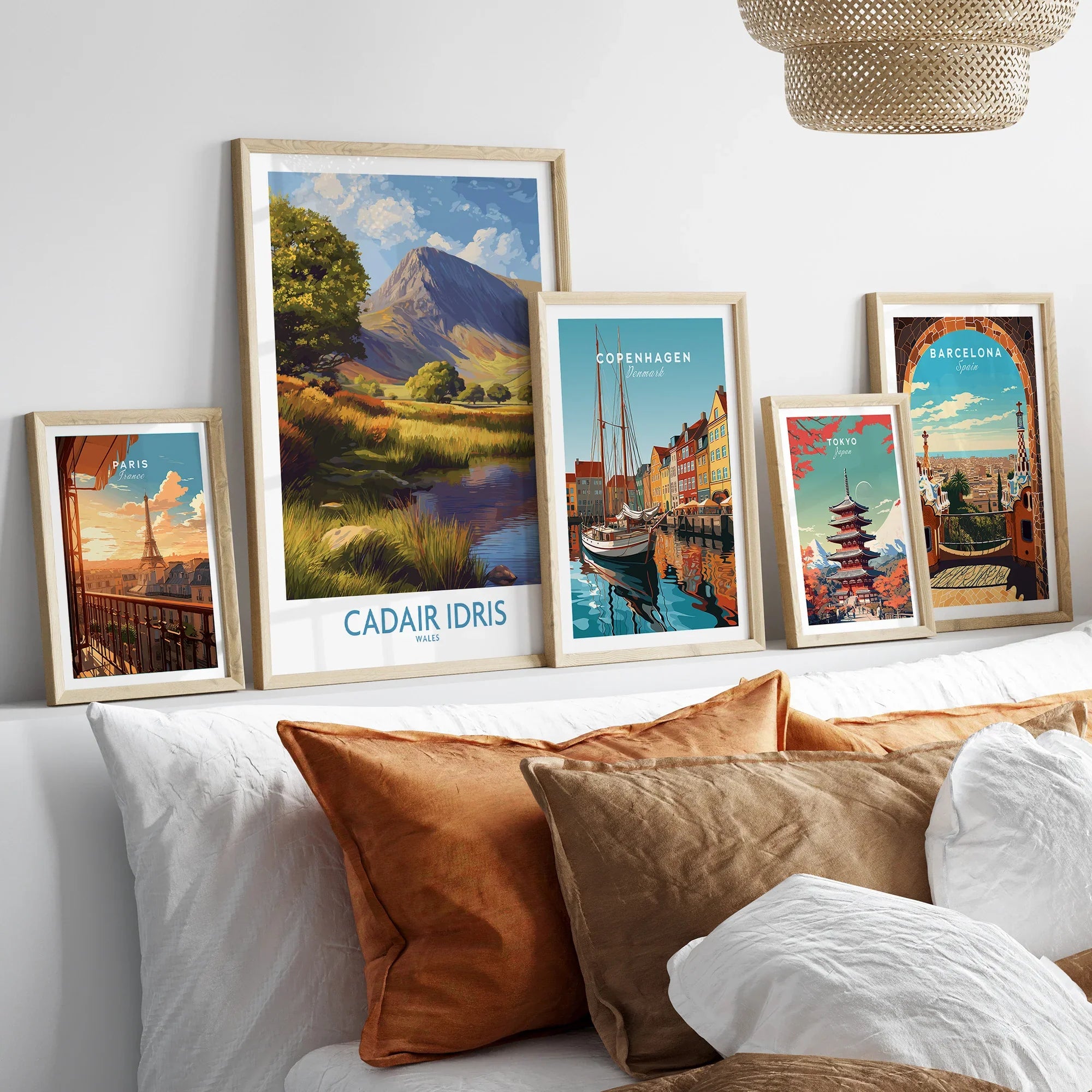 Set of five framed travel posters featuring Paris, Cadair Idris, Copenhagen, Tokyo, and Barcelona above bed with neutral pillows.
