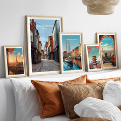 Set of five framed travel posters featuring Colchester, Copenhagen, Paris, Tokyo, and Barcelona above a bed with neutral wood frames and cozy pillows, perfect for home decor.