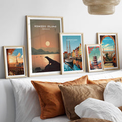 Set of five framed travel posters including Komodo Island, Copenhagen, Paris, Tokyo, and Barcelona above a bed with neutral pillows.