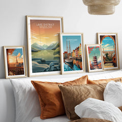 Set of five framed travel art prints featuring Paris, Lake District, Copenhagen, Tokyo, and Barcelona above a bed with natural wood frames.