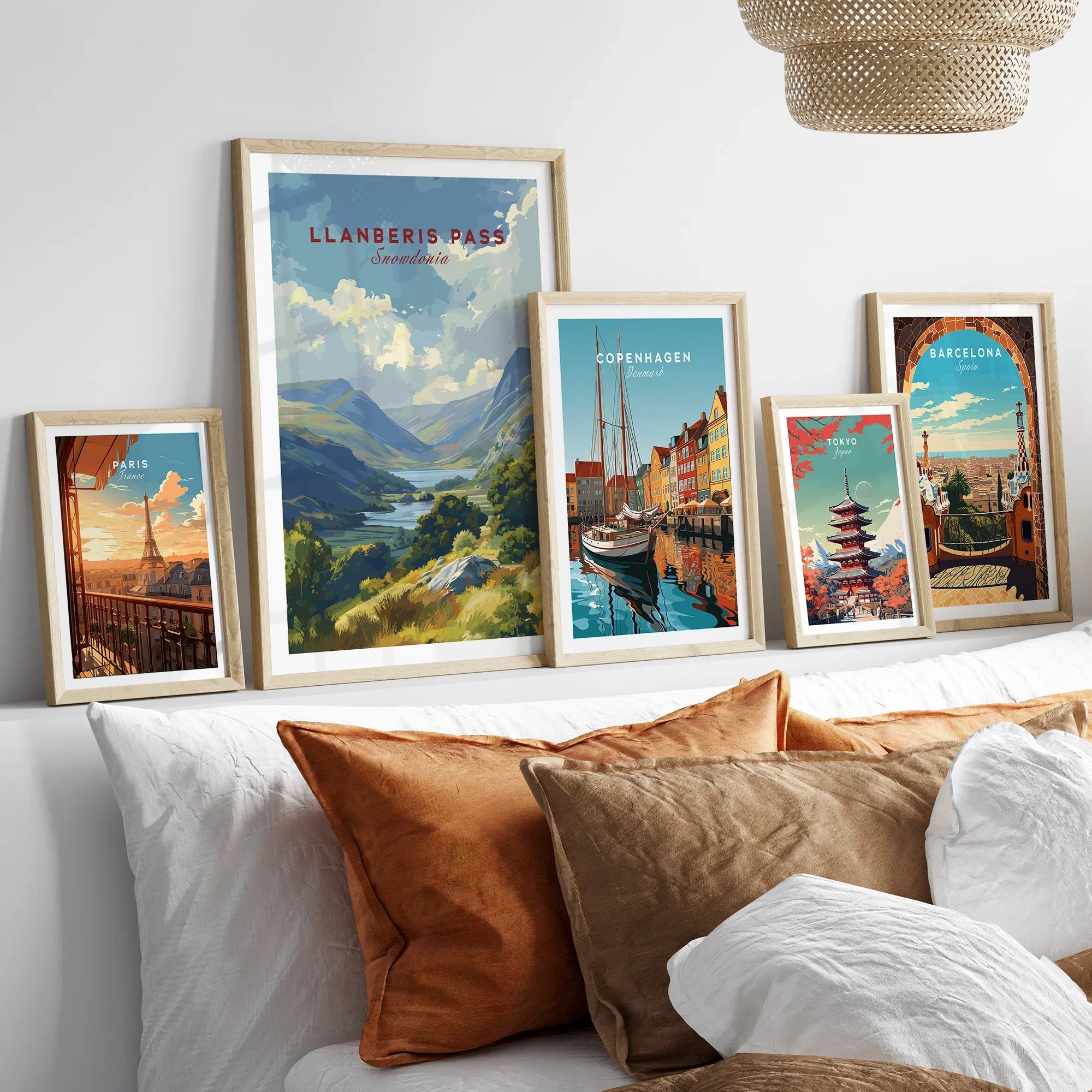 Set of five framed travel art prints featuring Paris, Snowdonia, Copenhagen, Tokyo, and Barcelona above a modern bed with earth-toned pillows.