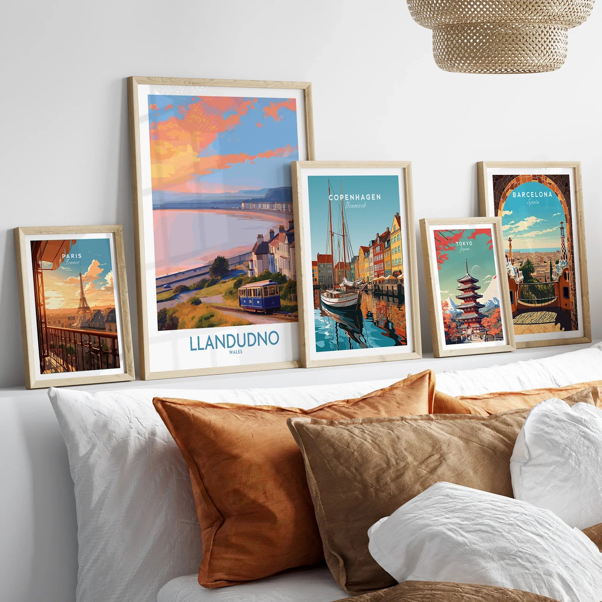 Set of five framed travel art prints above a bed, featuring landmarks from Paris, Llandudno, Copenhagen, Tokyo, and Barcelona in natural wood frames, perfect for wall decor.