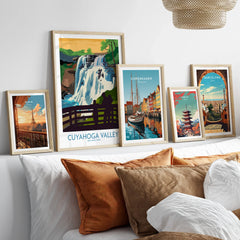 Set of five travel art prints featuring Paris, Cuyahoga Valley, Copenhagen, Tokyo, and Barcelona in natural wood frames above a bed.