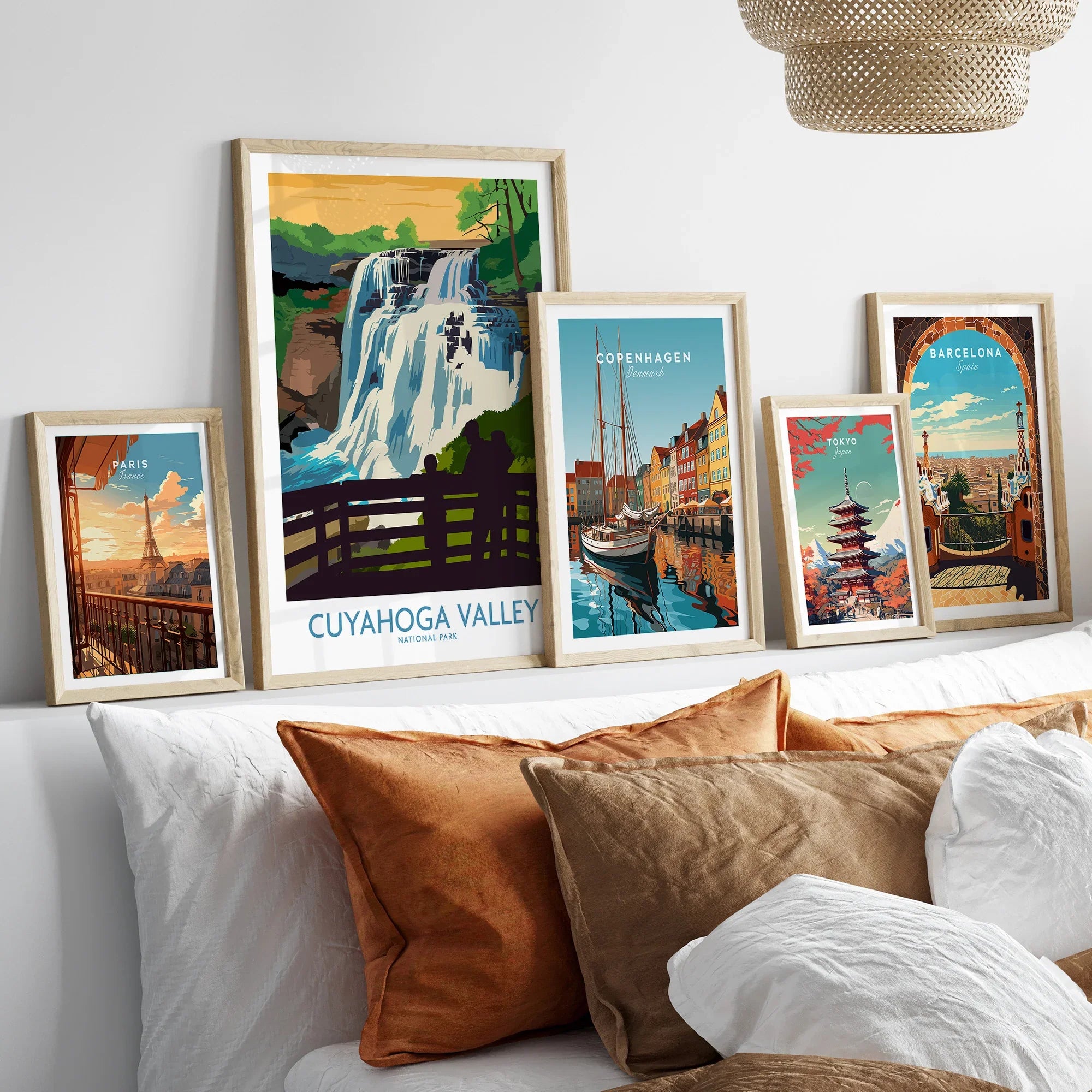 Set of five travel art prints featuring Paris, Cuyahoga Valley, Copenhagen, Tokyo, and Barcelona in natural wood frames above a bed.