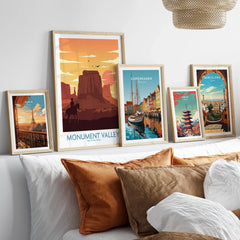 Set of five travel posters in natural wood frames above a bed, featuring Paris, Monument Valley, Copenhagen, Tokyo, and Barcelona wall art prints.