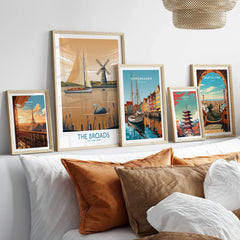 Set of five travel posters in natural wood frames above a bed, featuring Paris, The Broads, Copenhagen, Tokyo, and Barcelona wall art.