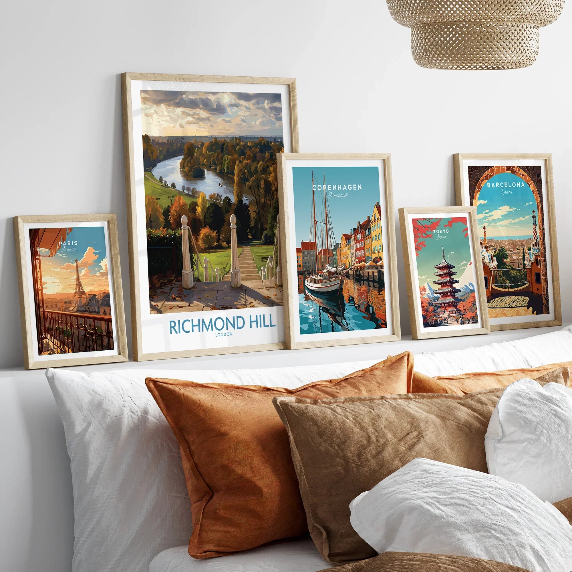 Set of five framed travel posters above a bed, featuring Paris, Richmond Hill, Copenhagen, Tokyo, and Barcelona artworks in natural wood frames.