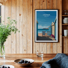 Cromer Travel Poster – Norfolk Beach Huts Art Print displayed in a rustic wooden setting with beach view.
