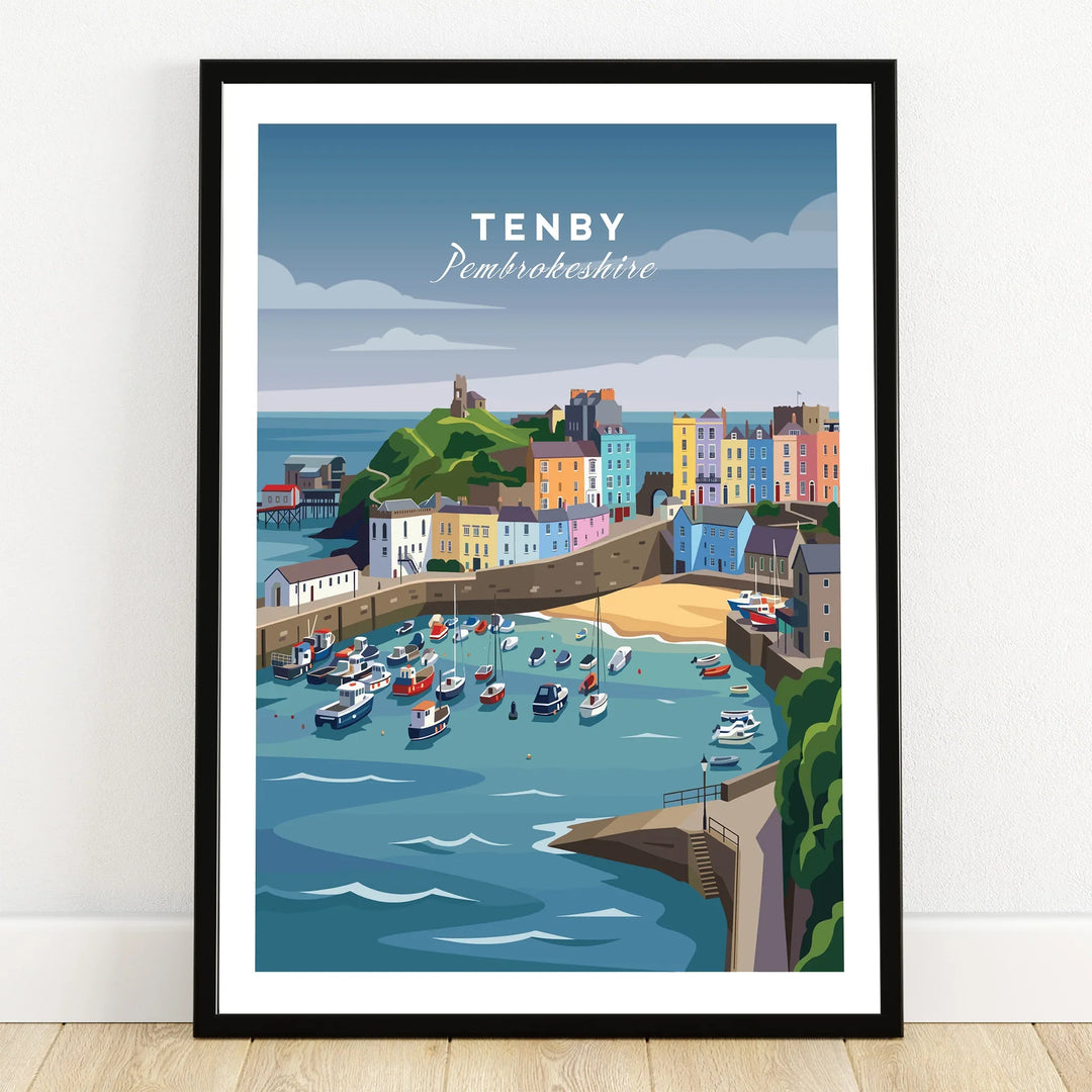 Tenby travel poster art print in black frame, featuring colorful harbor and boats, on wooden floor.