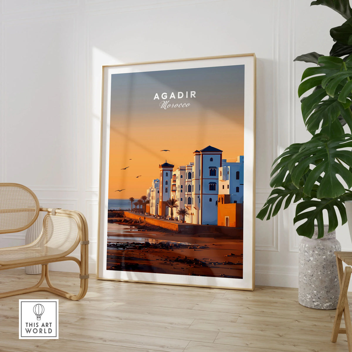 Agadir Travel Poster – Morocco Beach Print in a large natural wood frame leaning in a room with a rattan chair and monstera plant