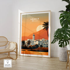 Agadir Travel Poster – Moroccan Mosque and Desert Sunset Art in a large natural wood frame leaning in a room with a rattan chair and monstera plant