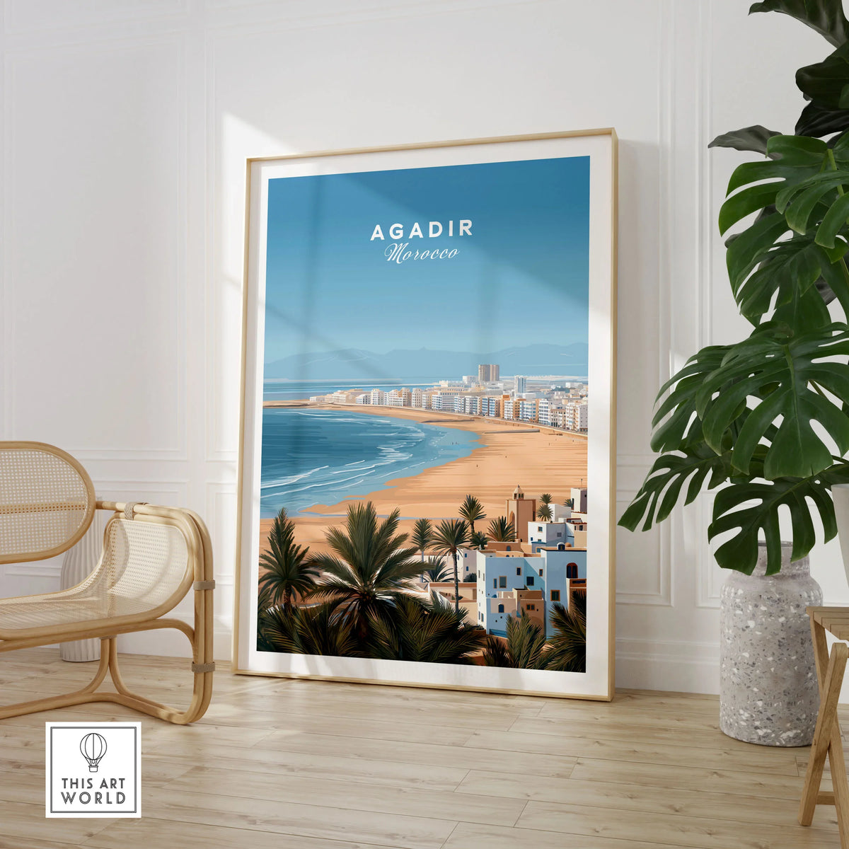 Agadir Morocco Poster – Travel Print in a large natural wood frame leaning in a room with a rattan chair and monstera plant