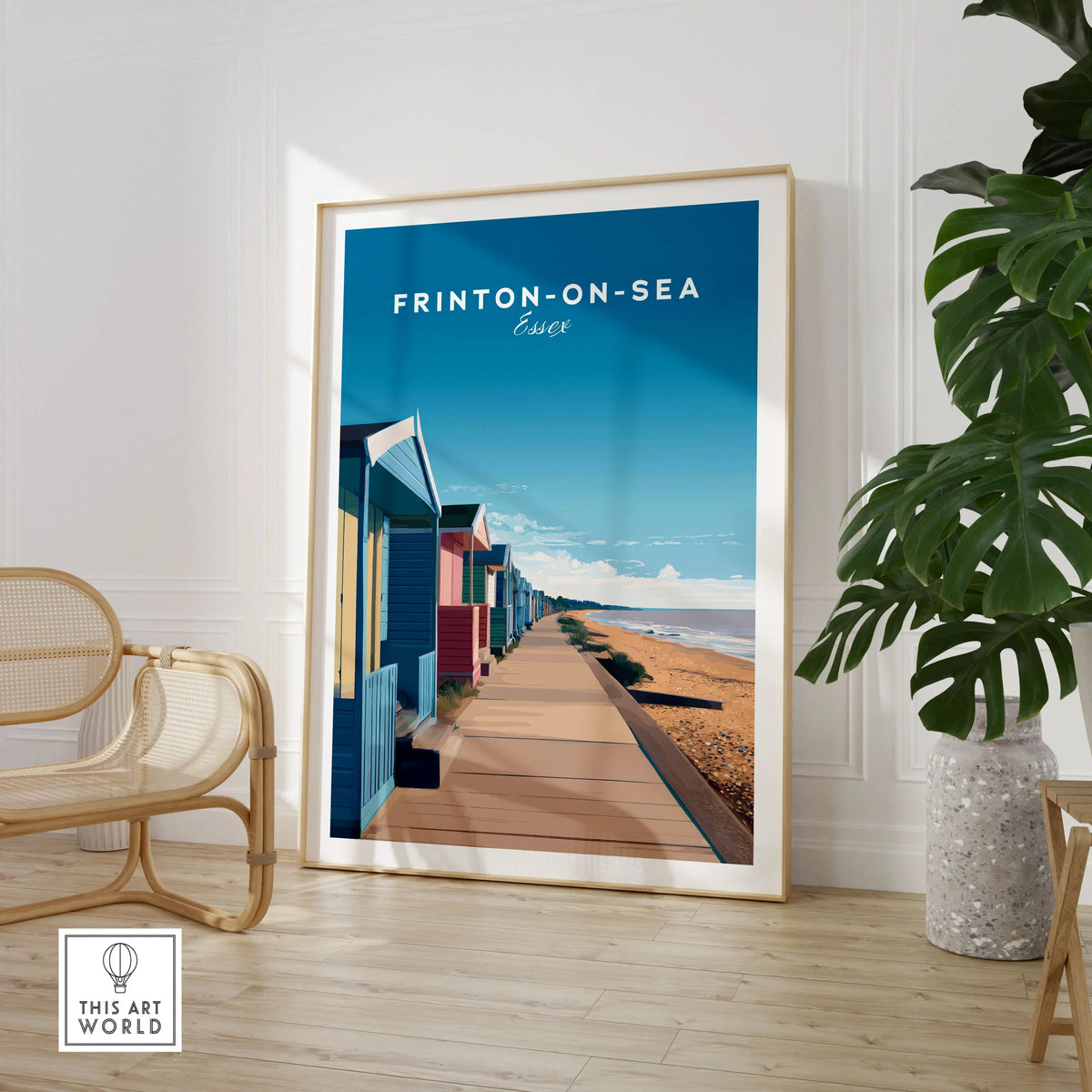 Frinton-on-Sea Travel Poster – Essex Coastline Beach Huts & Prom Art in a large natural wood frame leaning in a room with a rattan chair and monstera plant