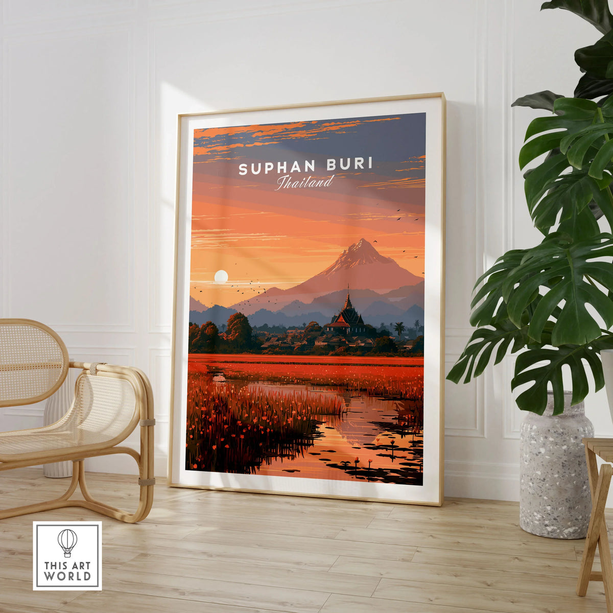Suphan Buri Thailand Lotus Lake Travel Poster — a lotus-covered lake and temple spire at sunset beneath a mountain peak, shown as large framed print room setting chair plant