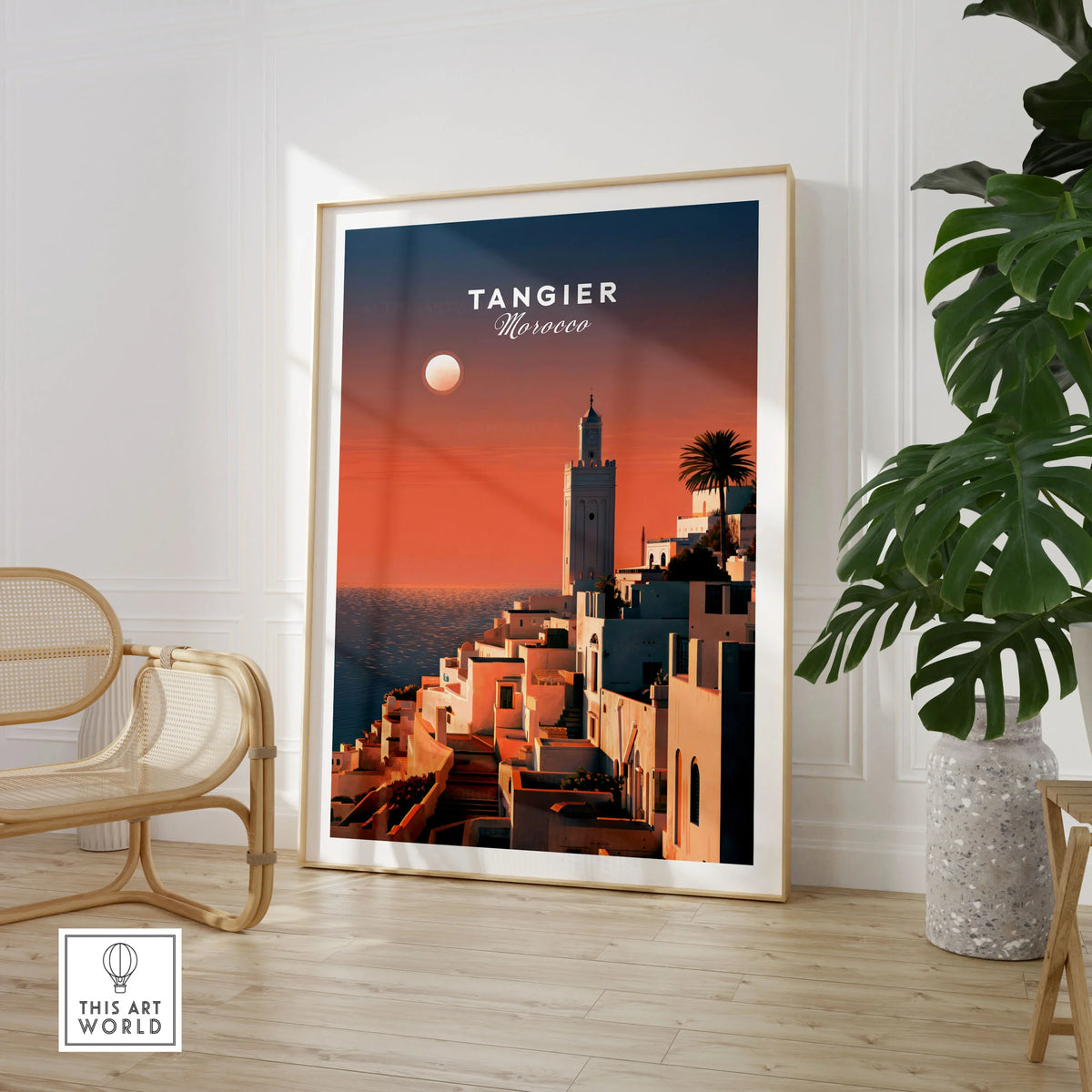 Tangier Morocco Golden Hour Travel Poster — a whitewashed hillside town and minaret glowing above the sea at sunset, shown as large framed print room setting chair plant