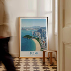 Framed Busan South Korea travel poster with city skyline and beach, leaning on floor in modern room