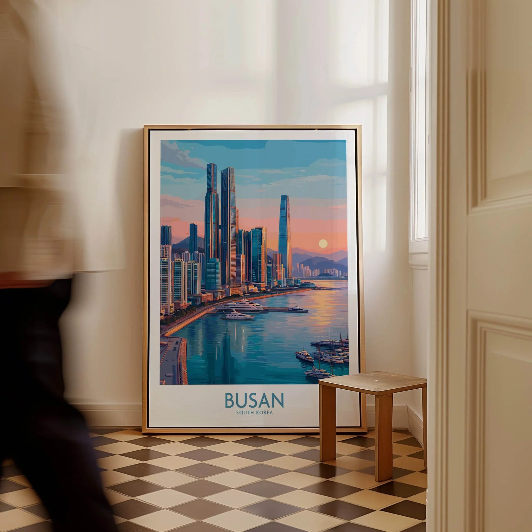 Framed Busan South Korea city skyline wall art on floor in sunlit hallway, man walking by