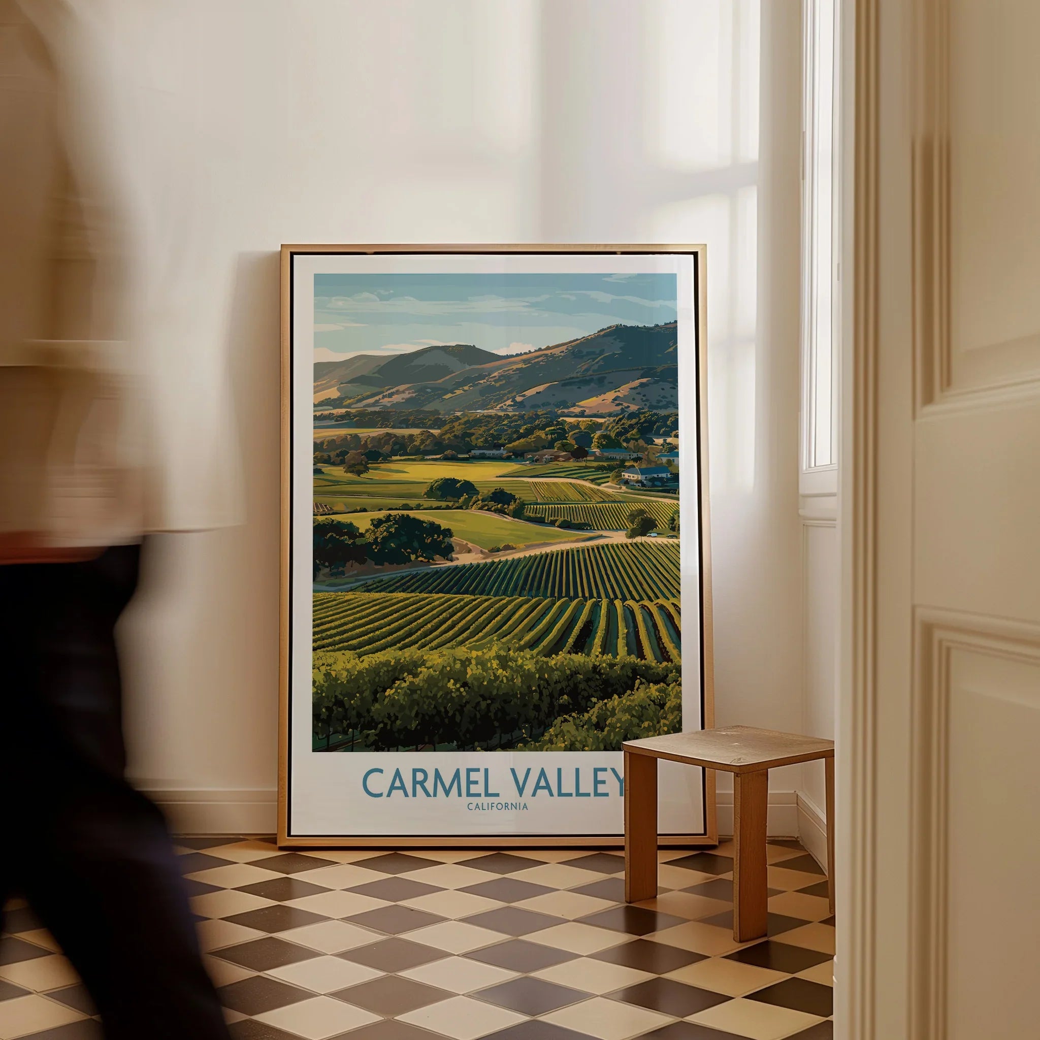 Framed Carmel Valley California travel poster leaning on floor in sunlit hallway, man walking by.