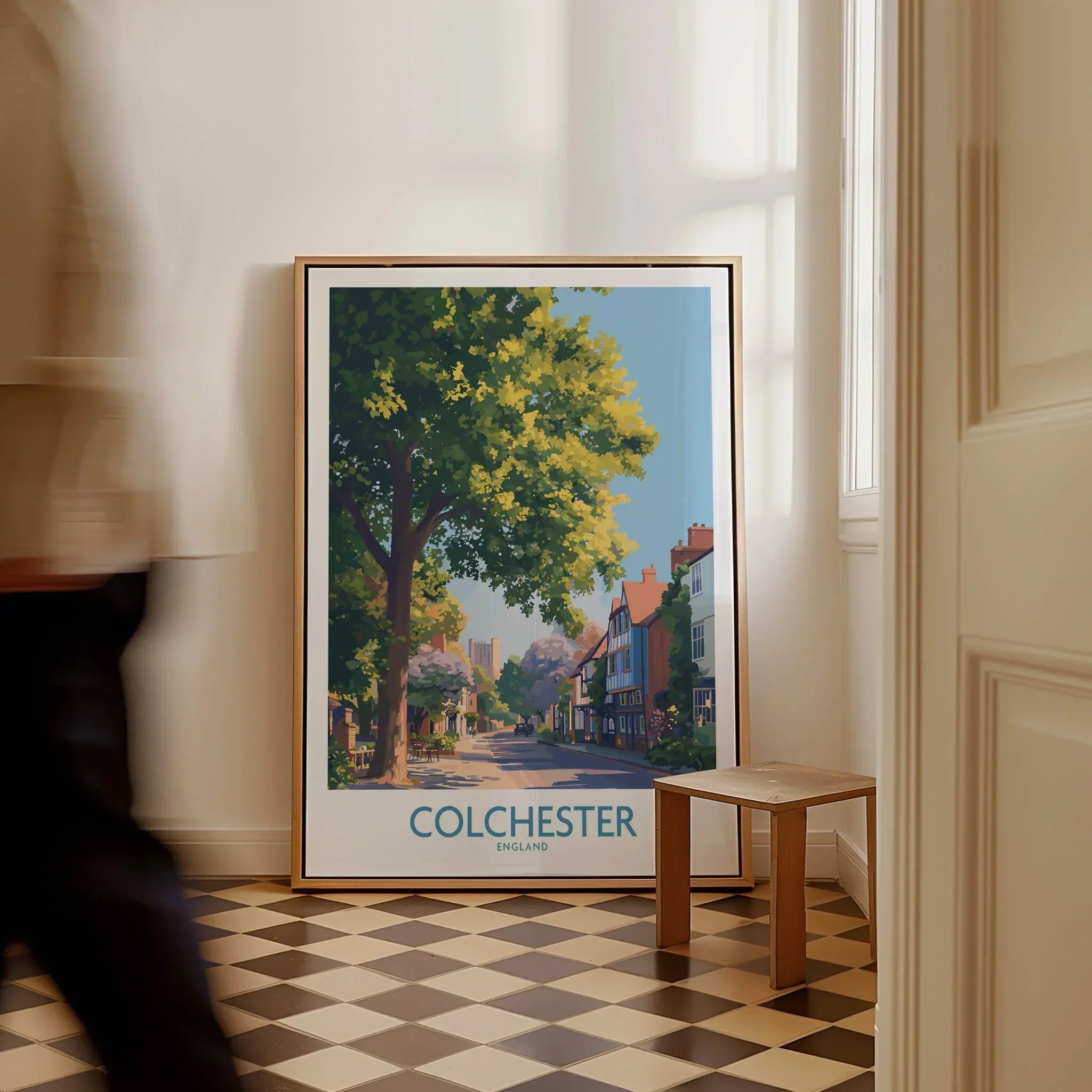 Framed Colchester England travel poster on checkered floor near window, man walking past.