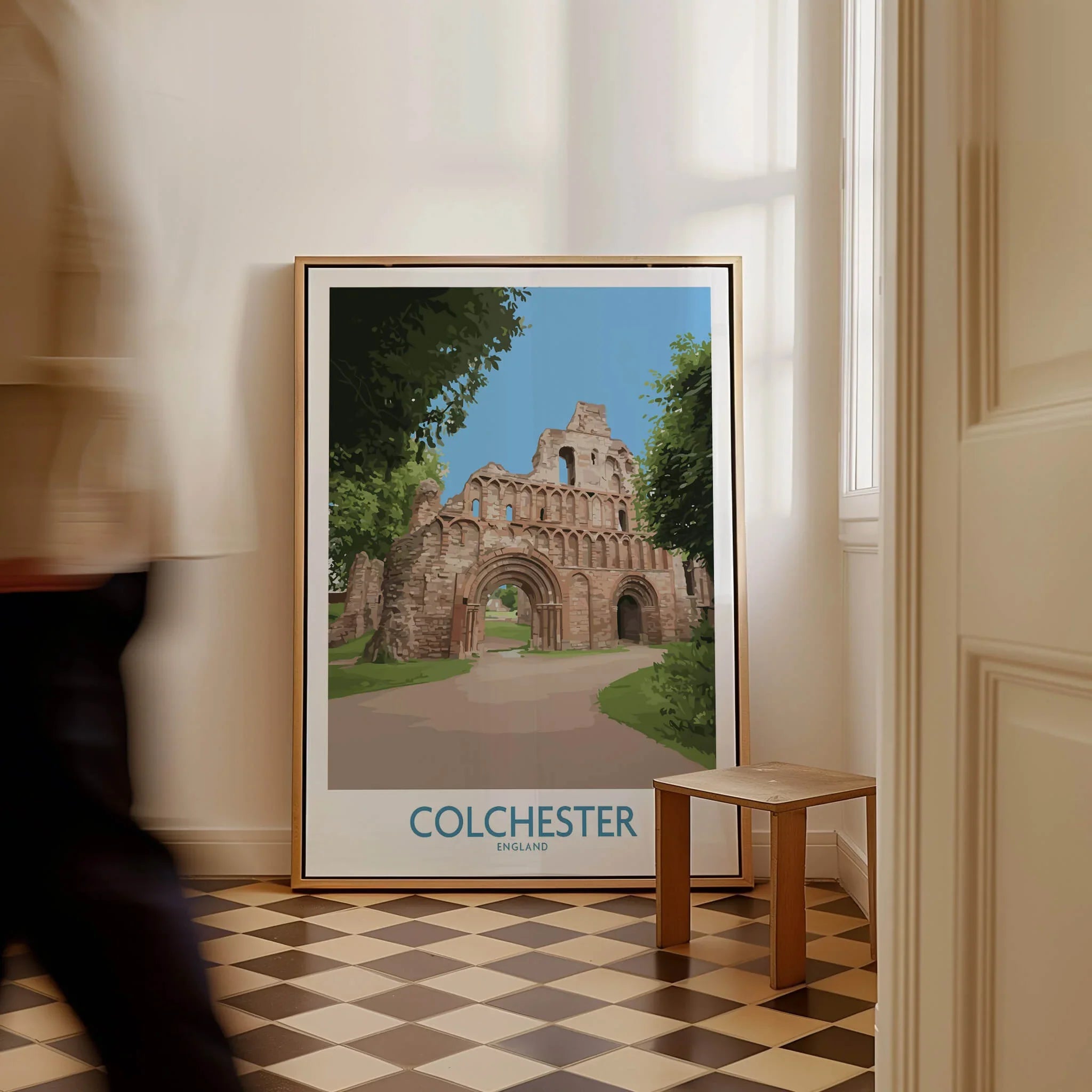 Framed Colchester travel poster with historic ruins, leaning on floor in modern home interior