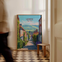 Framed Lewes England travel poster on floor in bright hallway, person walking past