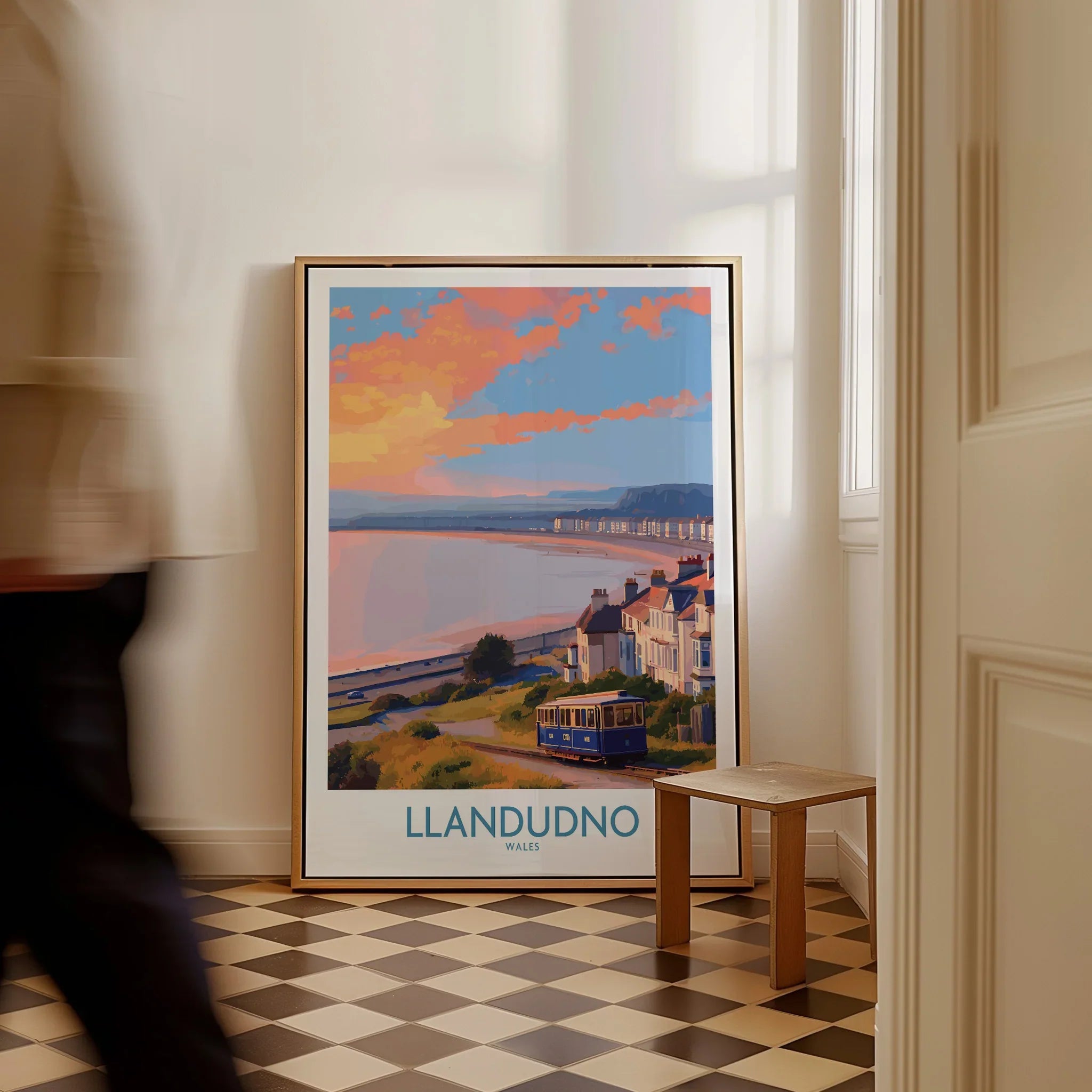 Framed Llandudno travel poster with coastal scene on floor, man walking past in home interior