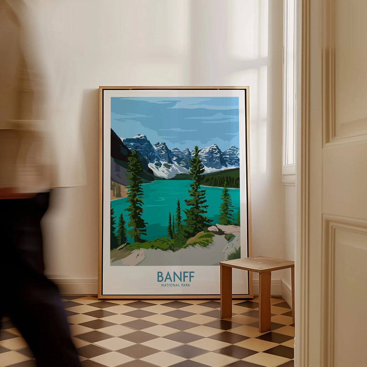 Framed Banff National Park travel poster on floor, man walking past in bright hallway
