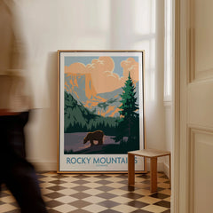 Framed Rocky Mountains Colorado travel poster with bear and sunset landscape on floor; man walking past
