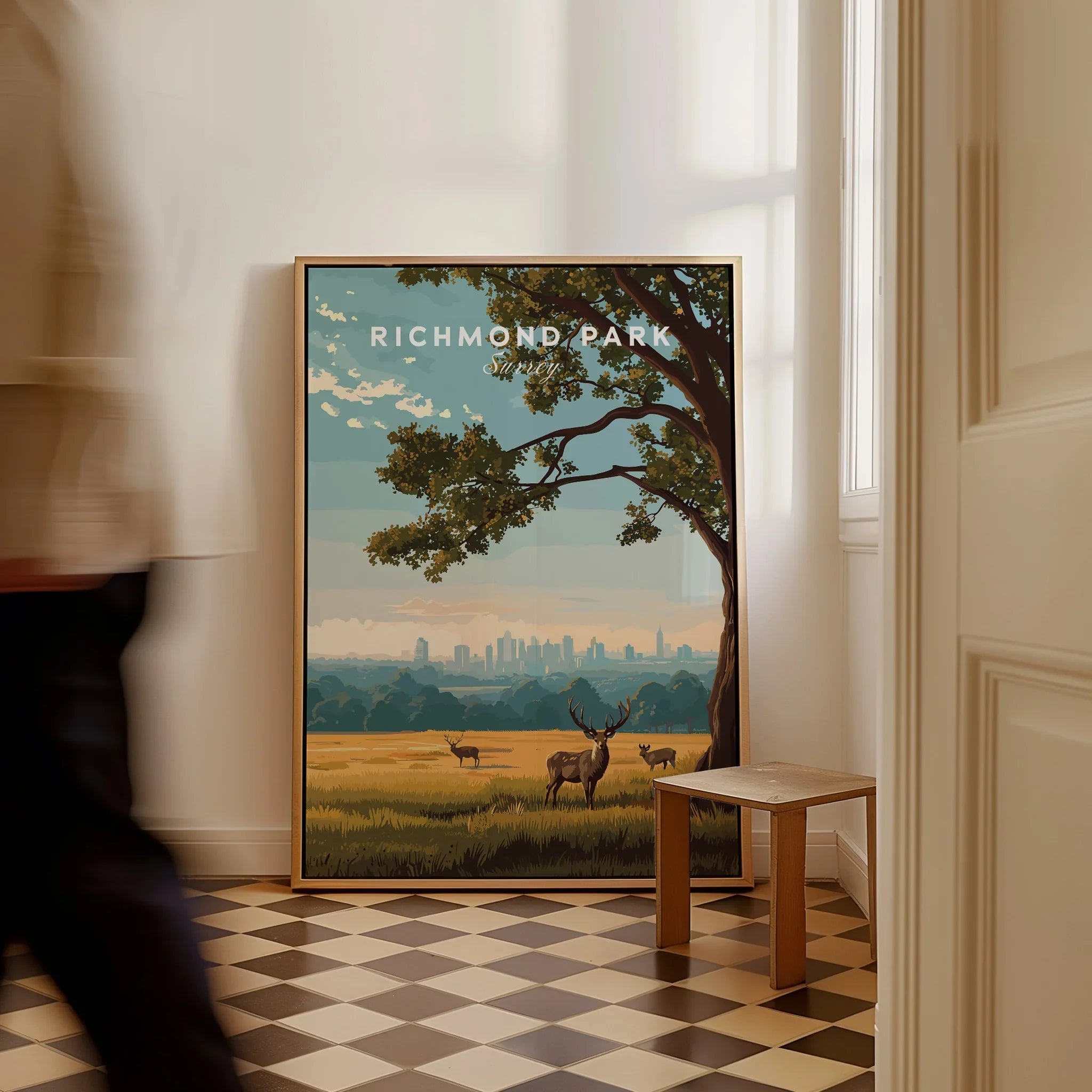 Framed Richmond Park travel poster with deer and London skyline leaning on floor, man walking past.
