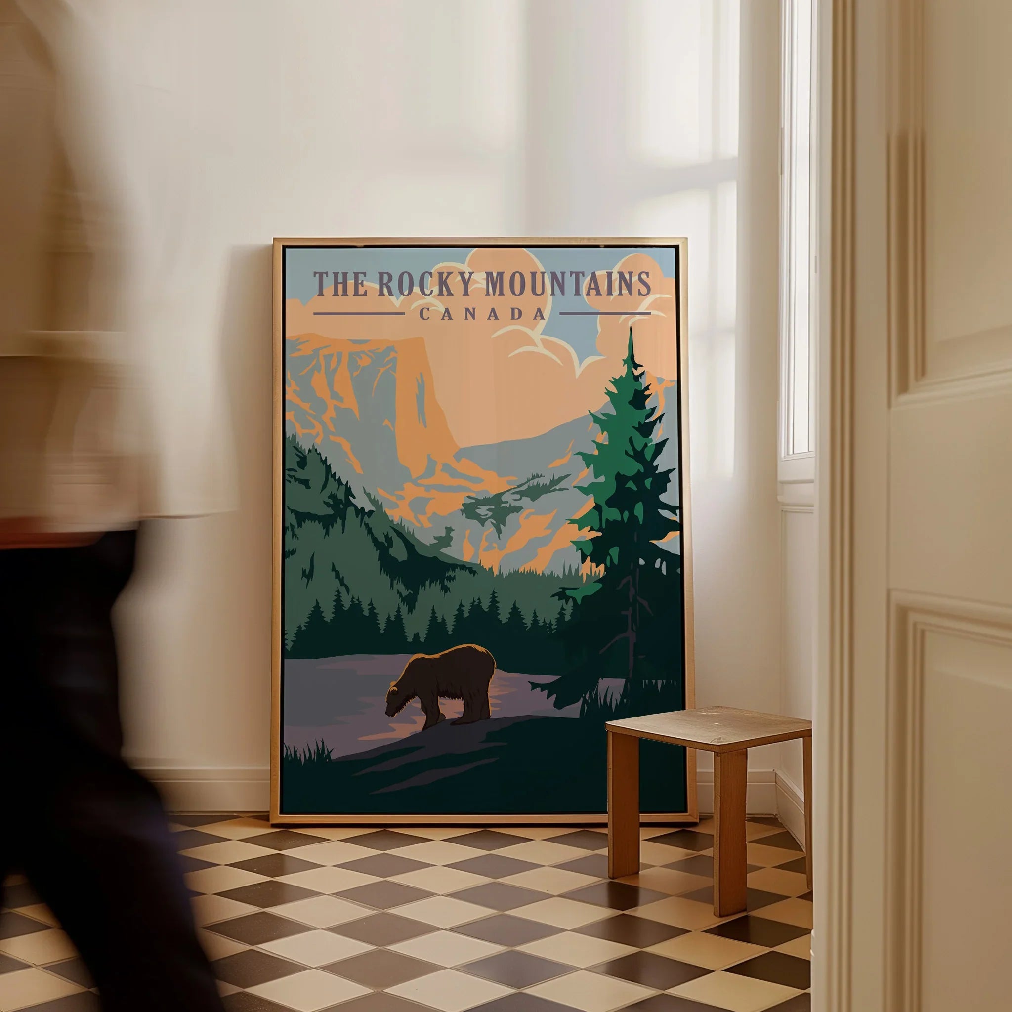 Framed Rocky Mountains Canada travel poster with bear and mountains, leaning on floor indoors.