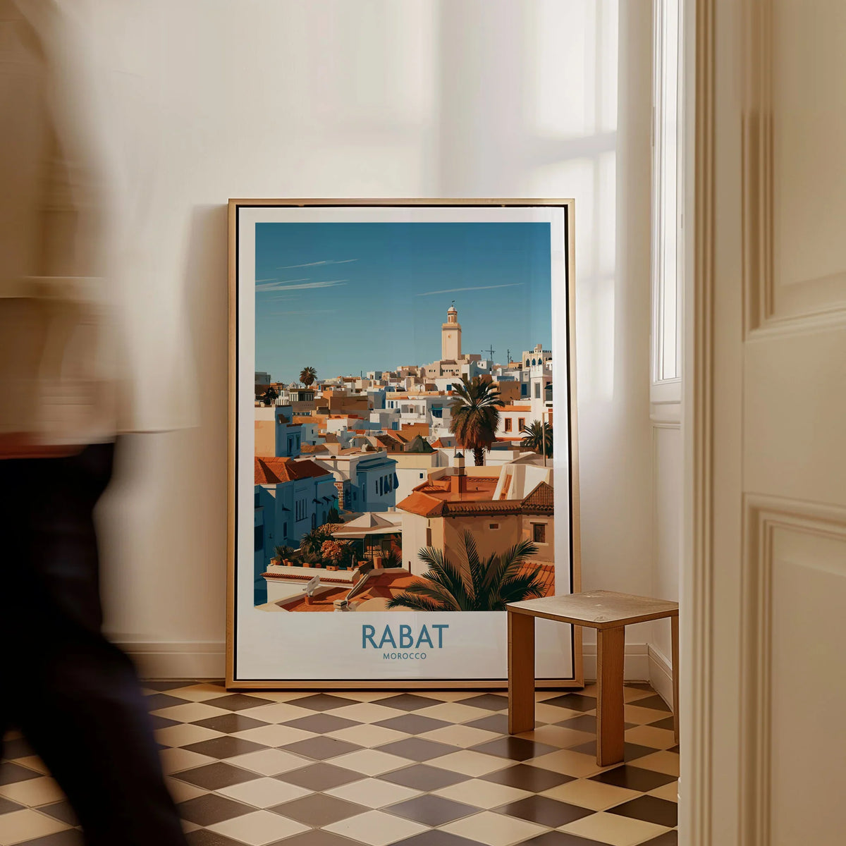 Rabat Morocco Minaret Skyline Wall Art — medina rooftops and a pale minaret in late afternoon sun, shown as framed canvas on floor man walking past
