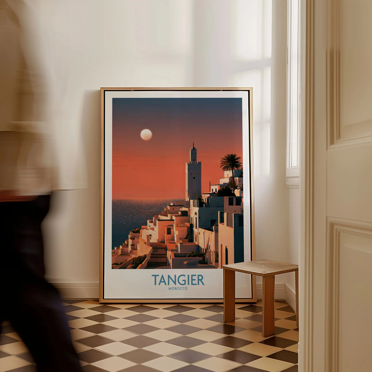 Tangier Morocco Coastal Sunset Wall Art — a whitewashed hillside town and minaret glowing above the sea at sunset, shown as framed canvas on floor man walking past