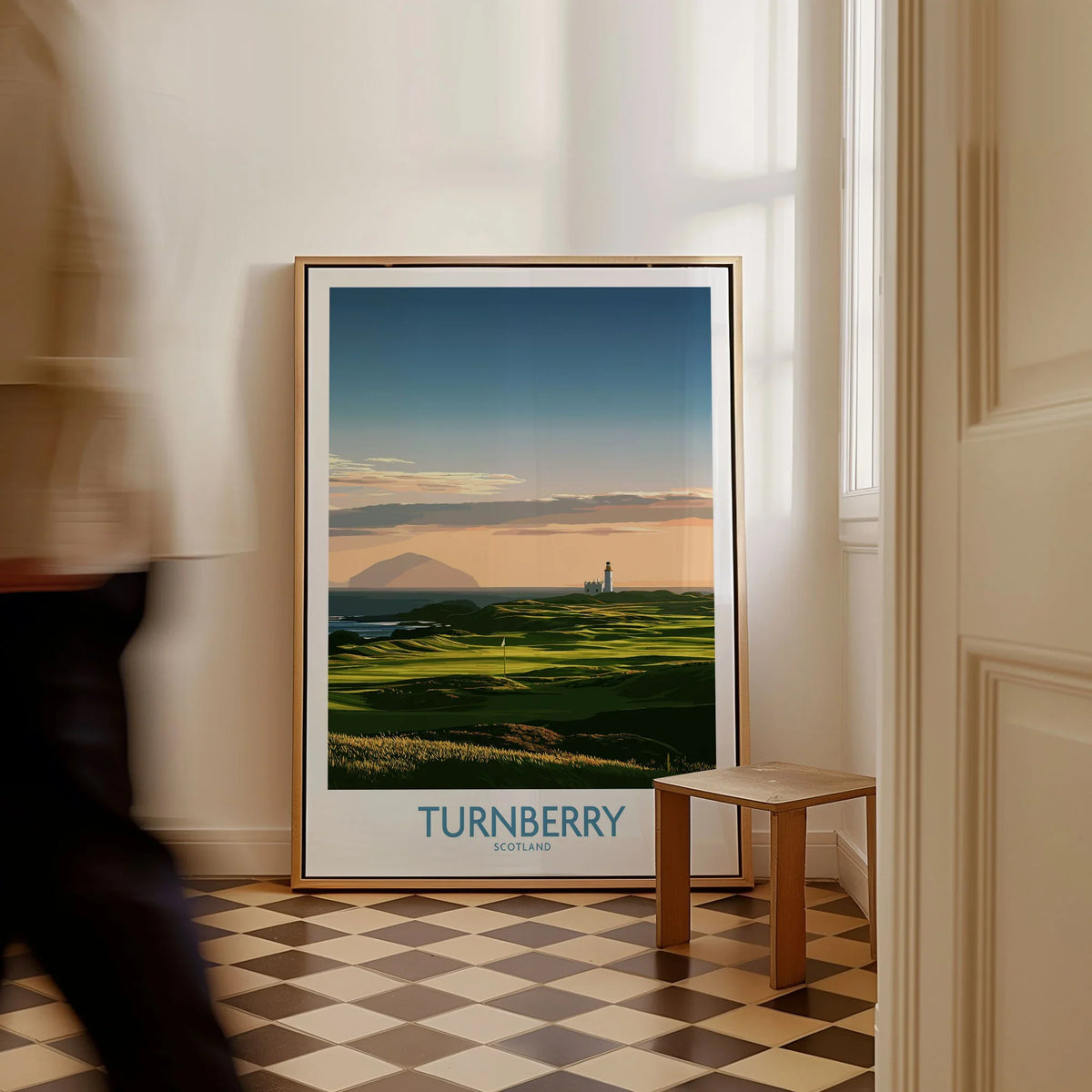 Turnberry Scotland Ayrshire Dusk Wall Art — Ailsa Craig and the white lighthouse across rolling coastal turf at dusk, shown as framed canvas on floor man walking past