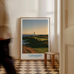 Turnberry Scotland Rocky Shore Wall Art — the Turnberry lighthouse above a rocky shore with the Arran hills beyond, shown as framed canvas on floor man walking past