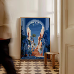 Rabat Morocco Blue Medina Travel Poster — a blue-washed medina lane and stone steps seen through a horseshoe arch, shown as framed canvas on floor man walking past