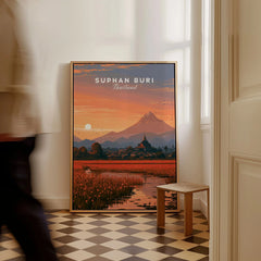 Suphan Buri Thailand Lotus Lake Travel Poster — a lotus-covered lake and temple spire at sunset beneath a mountain peak, shown as framed canvas on floor man walking past