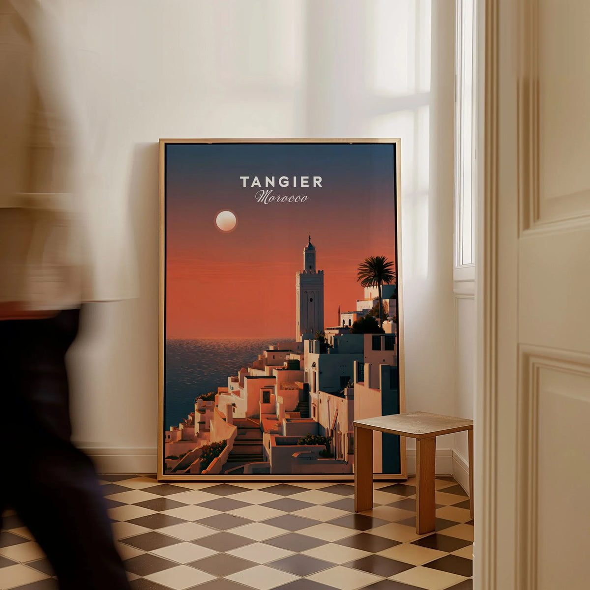 Tangier Morocco Golden Hour Travel Poster — a whitewashed hillside town and minaret glowing above the sea at sunset, shown as framed canvas on floor man walking past
