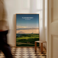 Turnberry Scotland Ailsa Craig Travel Poster — Ailsa Craig and the white lighthouse across rolling coastal turf at dusk, shown as framed canvas on floor man walking past
