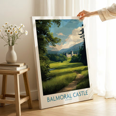 Hand holding Balmoral Castle travel poster in cozy room with wooden stool and flowers.