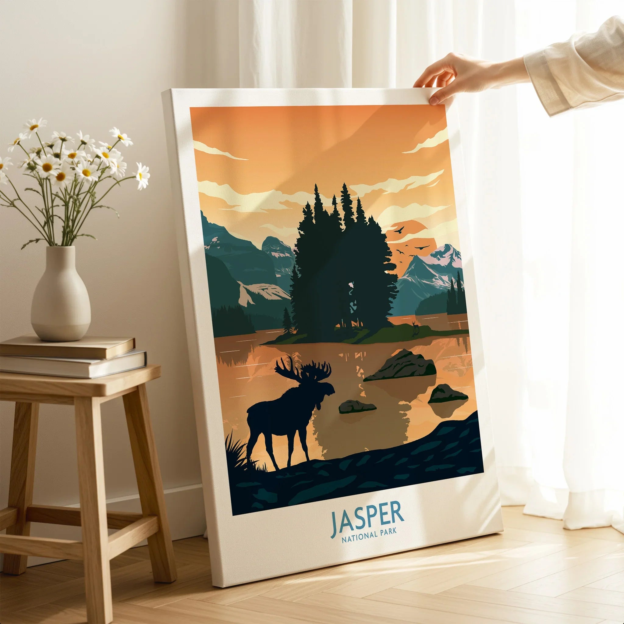 Hand holding Jasper National Park canvas print with moose and mountain lake artwork in cozy room.