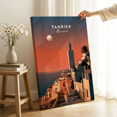 Tangier Morocco Golden Hour Travel Poster — a whitewashed hillside town and minaret glowing above the sea at sunset, shown as canvas print mirror edge hand holding room setting